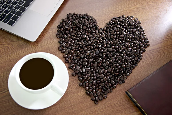Coffee beans arranged shaped into a heart near white coffee cup. — Stock Photo, Image