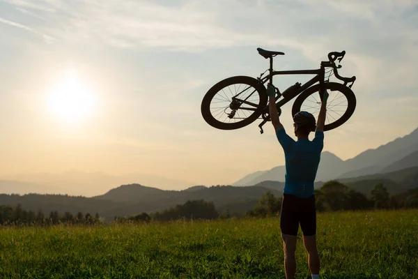 Ciclista subindo uma bicicleta acima de sua cabeça e olhando para o pôr do sol — Fotografia de Stock