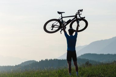 Bisikletçi kafasının üstünde bir bisiklet yükseliyor ve gün batımını seyrediyor.