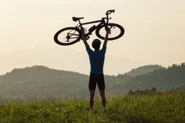 Bisikletçi kafasının üstünde bir bisiklet yükseliyor ve gün batımını seyrediyor.