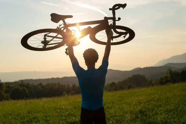 Ciclista subindo uma bicicleta acima de sua cabeça e olhando para o pôr do sol — Fotografia de Stock