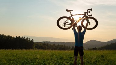Bisikletçi kafasının üstünde bir bisiklet yükseliyor ve gün batımını seyrediyor.