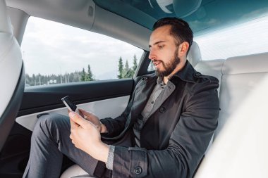 Beyaz tenli başarılı genç adam, bir arabanın arka koltuğunda otururken ciddi ciddi akıllı telefondan konuşuyor.