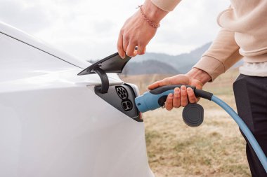 Erkek, arka plandaki elektrikli araba soketine şarj aleti yerleştiriyor.