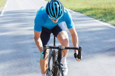 Caucasian man in professional road cycling clothes, a blue jersey, with a helmet, sunglasses, and gloves, riding uphill on an asphalt road