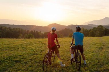 Profesyonel bisikletçi çift yeşil bir çayırda mola veriyor ve muhteşem dağ günbatımını seyrediyor..