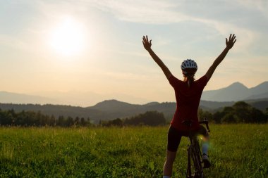 Dişi bisiklet yarışçısı Dağ manzarasında açık bir yolda pedal çeviriyor, bir arabanın yanından geçiyor ve zafer pozuyla kollarını kaldırıyor, geniş açıyla..