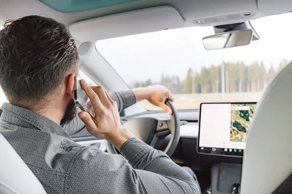 Male driver at the wheel of a electric car, driving and talking on smartphone, rear view.