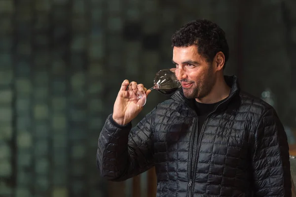 Man tasting red wine in a winery barrel cellar, holding a wine glass, swirling it, smelling, and sipping wine, delighted with wine taste and flavor.