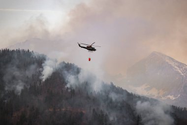 Firefighting helicopter with a bucket suspended on a cable, collecting water from a mountain lake to extinguish the flame in a forest