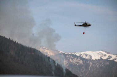Firefighting helicopter with a bucket suspended on a cable, collecting water from a mountain lake to extinguish the flame in a forest