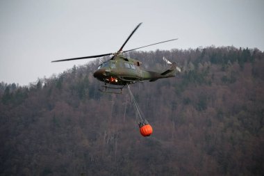Firefighting helicopter with a bucket suspended on a cable, collecting water from a mountain lake to extinguish the flame in a forest