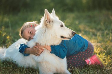 Caucasian sweet little girl hugging and cuddling a beautiful white dog, close up shot. Animals, dogs, love, and friendship concept.