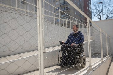 Man on electric wheelchair moving along concrete accessibility pathway near the building. Concept of wheelchair access facilitates.