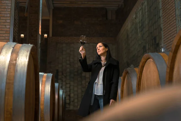Woman in the wine cellar with barrels in background drinking and tasting wine.