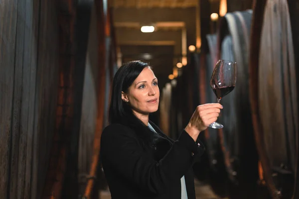 Woman in the wine cellar with barrels in background drinking and tasting wine.