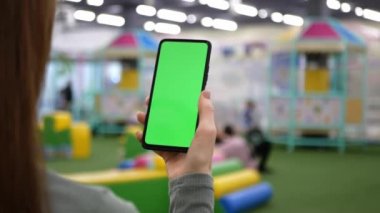 Genç anne yeşil ekranlı akıllı telefon kullanıyor. Halka açık yerlerde internet sörfü kavramı. Çocuklar eğlence ya da oyun merkezinde beklerken kadın yeşil model ekrana vuruyor.
