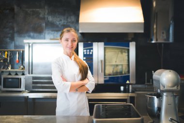 Portrait of confident and smiling young woman chef dressed in white uniform, professional kitchen are on background. Restaurant  kitchen