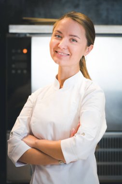 Portrait of confident and smiling young woman chef dressed in white uniform, professional kitchen are on background. Restaurant  kitchen