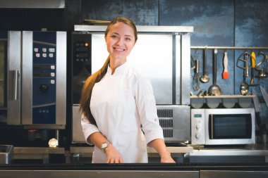 Portrait of confident and smiling young woman chef dressed in white uniform, professional kitchen are on background. Restaurant  kitchen
