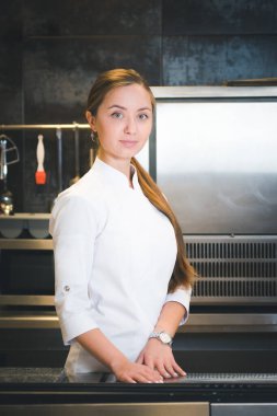 Portrait of confident and smiling young woman chef dressed in white uniform, professional kitchen are on background. Restaurant  kitchen
