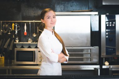 Portrait of confident and smiling young woman chef dressed in white uniform, professional kitchen are on background. Restaurant  kitchen