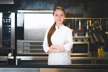 Portrait of confident and smiling young woman chef dressed in white uniform, professional kitchen are on background. Restaurant  kitchen