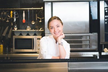 Portrait of confident and smiling young woman chef dressed in white uniform, professional kitchen are on background. Restaurant  kitchen