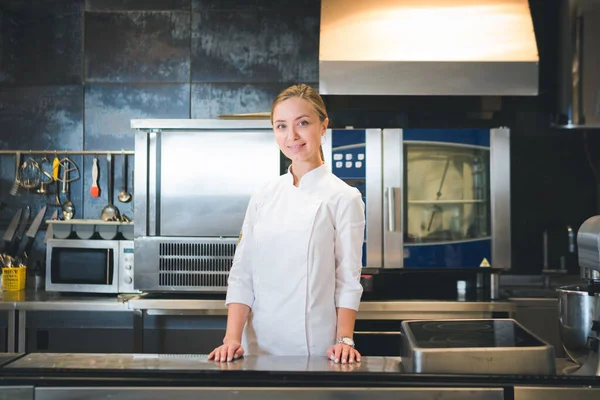 Portrait of confident and smiling young woman chef dressed in white uniform, professional kitchen are on background. Restaurant  kitchen