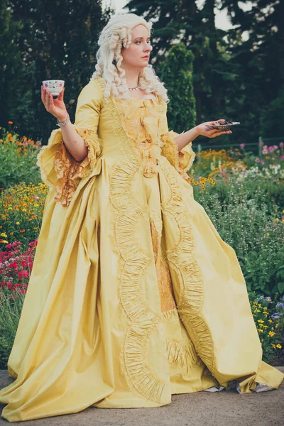 Portrait of blonde woman dressed in historical Baroque clothes with old fashion hairstyle, outdoors. Golden color dress. Luxurious medieval dress