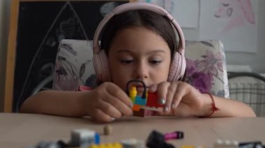 Şirin küçük kız ahşap masada oturuyor ve renkli lego yapı taşlarıyla oynuyor. Küçük kız ritim tutuyor ve kulaklıkla müzik dinleyerek eğleniyor..