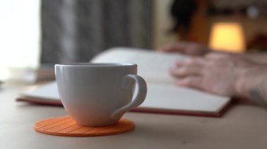 Çay ya da kahve içen adam sıcak bir fincan sıcak içecekten bir yudum alır ve kitabın sayfalarını çevirir. Pencereden gelen ışık iş yüzeyine düşer. Bulanık arkaplan
