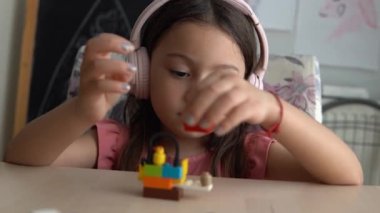 Şirin küçük kız ahşap masada oturuyor ve renkli lego yapı taşlarıyla oynuyor. Küçük kız ritim tutuyor ve kulaklıkla müzik dinleyerek eğleniyor..