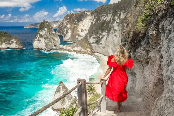 印度尼西亚巴厘岛努沙佩尼达市，一名身穿红色连衣裙站在通往钻石海滩的楼梯上的后视镜女子. 图库图片