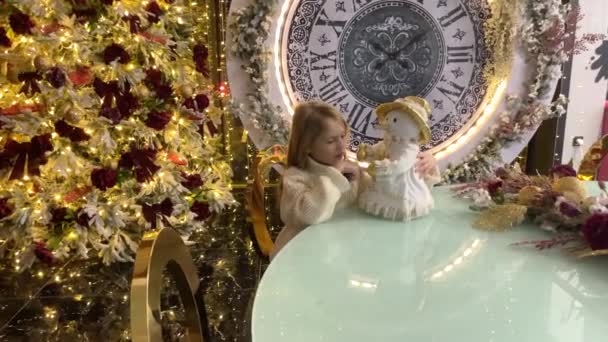 Une petite fille avec un bonhomme de neige près d'un sapin de Noël avec une grande horloge. Le concept de nouvelle année et de Noël. — Video