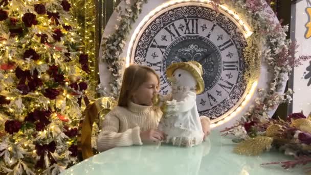 Una niña juega con un muñeco de nieve cerca de un árbol de Navidad con un reloj grande. El concepto de año nuevo y Navidad. — Vídeo de stock