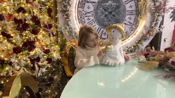 Una niña juega con un muñeco de nieve cerca de un árbol de Navidad con un reloj grande. El concepto de año nuevo y Navidad. — Vídeos de Stock