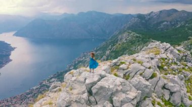 Karadağ 'daki Kotor Körfezi' ne bakan bir dağın tepesinde mavi elbiseli genç bir kız duruyor. Hava aracı görüntüsü 4K. Yaşam tarzı turizm ve güzellik konsepti.
