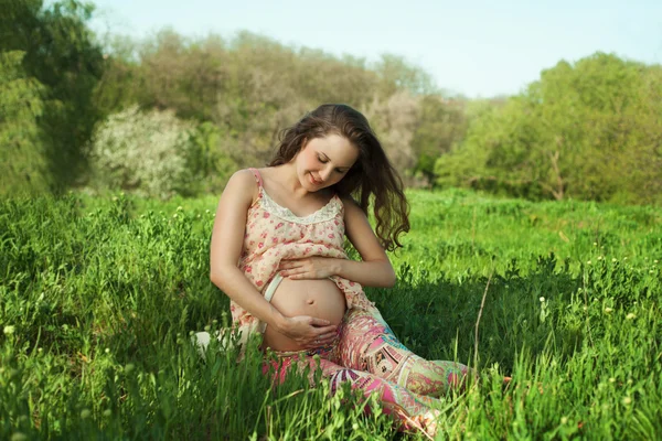 Mujer embarazada sentada en la hierba mirando su barriga mientras Imagen de archivo