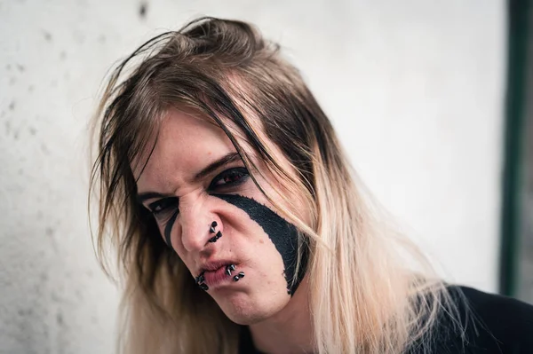 Head shot  of an attractive emo punk young man looking with anger and strong grimace  directly in camera. Close up of a pierced and face painted, long hair guy in his late 20s.