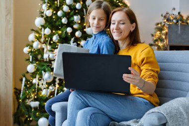 Noel çevrimiçi aile selamları. Gülümseyen Avrupalı anne ve kız, arkadaşlarına ve ebeveynlerine video araması yapmak için dizüstü bilgisayar kullanıyor.