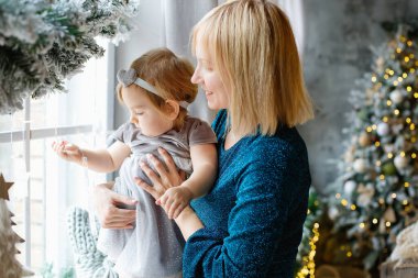 Anne ve bebek pencerede oturuyor. Noel süsleri. Noel 'de, bir mucize bekliyor. Pencereden dışarı bakan çocuklu bir kadın..