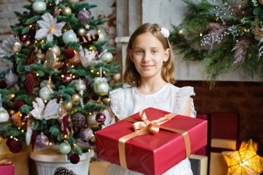 Seçici odaklanma. Kırmızı hediye kutusu olan beyaz elbiseli bir kız Noel ağacı ve şömineyle süslenmiş bir odada..
