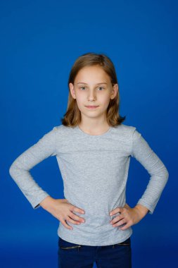 Bir kız mavi arka planda gri uzun kollu poz veriyor. Kameraya bakarken ellerini beline koy..