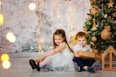 İki neşeli giyimli çocuk, bir oğlan ve bir kız, Noel için süslenmiş bir odada oturuyorlar, arka planda çelenklerin ışıklarına karşı. Erkek ve kız kardeşlerin bayram hediyeleri var..