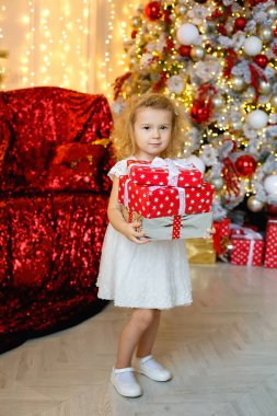 Beyaz elbiseli küçük kıvırcık sarışın bir kızın büyük bir portresi. Elinde hediyeler, Noel ağacı ve çelenk ışıkları olan bir odada..