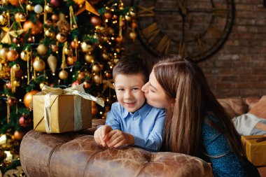Anne ve oğul, altın toplar ve çelenklerle süslenmiş bir Noel ağacının yanında kucaklaşıyor. Anne çocuğu yanağından öper..
