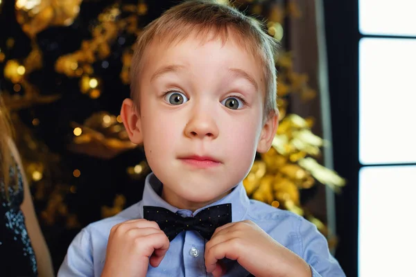 A boy in smart clothes on the background of a Christmas tree looks at the camera and adjusts his bow tie.