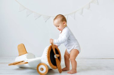 Beyaz elbiseli küçük bir çocuk içeride oyuncak bir uçakla eğleniyor. Beyaz zeminde bir çocuk oynuyor..
