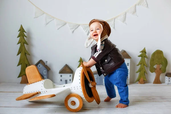 A happy one-year-old boy in a helmet and a pilot\'s jacket stands near a wooden plane. Portrait in the studio of a child aviator.